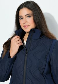 Ung kvinna med långt mörkt hår som drar upp en marinblå vadderad jacka mot en vit bakgrund.