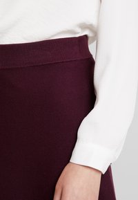 Burgundy kjol med ribbad midja, ihopparad med en vit långärmad skjorta med en strukturerad krage. Slät textur och figurnära design.