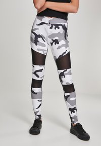 Camo mintázatú legginsz fehér, szürke és fekete sz�ínben, átlátszó hálós betéttel. Fitt fazon, rugalmas anyaggal a kényelem érdekében. Fekete sportcipővel viselve.