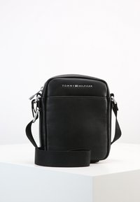 Black leather shoulder bag with a rectangular shape, featuring a front logo and an adjustable fabric strap. Smooth texture, minimal design.