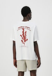 Camiseta blanca de manga corta con un gráfico floral rojo y texto en la parte posterior, hecha de algodón y con un corte relajado. Los pantalones cortos son de lino beige.
