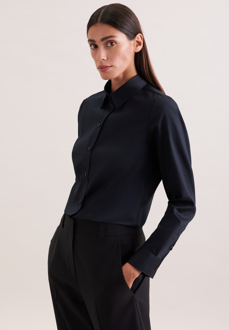 Femme aux cheveux longs et foncés portant une chemise noire ajustée à boutons et un pantalon noir, debout avec une main dans la poche, devant un fond clair.