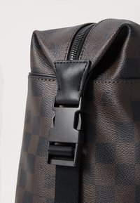 Brown patterned backpack featuring a textured surface, black plastic buckle, and zip closure at the top.