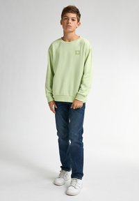 Lichtgroene sweater met een ronde hals, lange mouwen, een rechthoekige logopatch, gecombineerd met donkerblauwe jeans en witte sneakers.