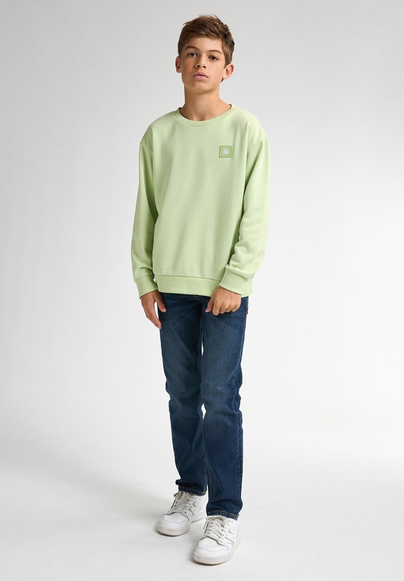Lichtgroene sweater met een ronde hals, lange mouwen, een rechthoekige logopatch, gecombineerd met donkerblauwe jeans en witte sneakers.