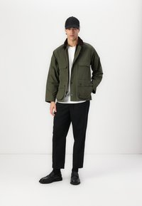 Olive green, zippered jacket with two front flap pockets, brown collar, worn over a white shirt, paired with black tapered trousers and shoes.