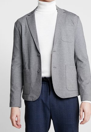 Blazer grigio strutturato con due bottoni, rever a scollo e tasche frontali. Indossato sopra un dolcevita bianco e abbinato a pantaloni a quadri.