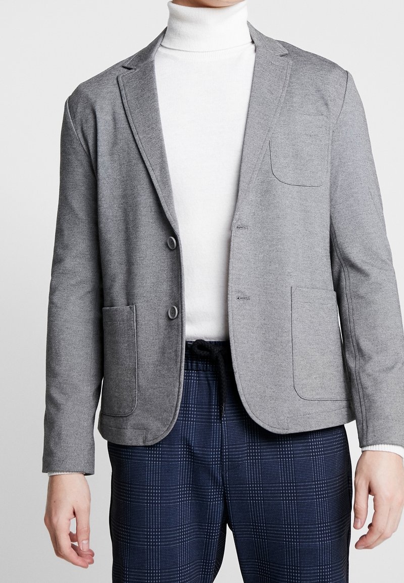 Blazer grigio strutturato con due bottoni, rever a scollo e tasche frontali. Indossato sopra un dolcevita bianco e abbinato a pantaloni a quadri.