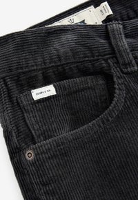 Sorte kordfløjlsbukser med en frontlomme, hvor der står "SUPPLY CO." på et label. Teksturen er ribbet med en subtil glans.