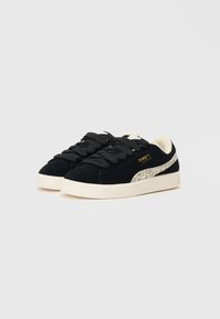 Juodi zomšiniai sportbačiai su tekstūruota balta guma, turi tamsius raiščius ir auksinį logotipą, papildyti kontrastinga šviesia akcento spalva.