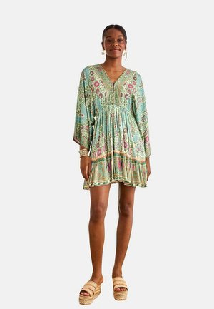 Mujer que lleva un vestido corto de manga larga con estampado floral en tonos verdes y rosas, combinado con sandalias con plataforma beige y joyas doradas.