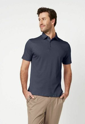 Homme portant un polo bleu marine à manches courtes et un pantalon beige, debout les mains dans les poches, regardant sur le côté sur fond blanc.