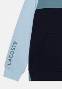 Sweatshirt en color-block présentant des panneaux bleu clair, turquoise et marine. Fabriqué en tissu doux avec des poignets côtelés et une poche avant. "LACOSTE" imprimé sur la manche.