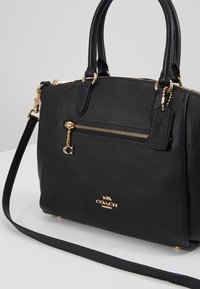 Borsa a mano in pelle nera con hardware dorato, esterno testurizzato, due manici, tracolla rimovibile e tasca frontale con zip. Logo Coach visibile.