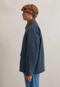 Plava vanjska jakna s smeđim ovratnikom, džepovima i slobodnim krojem. Tkanina izgleda glatko s blagim sjajem.