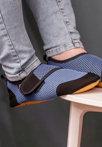 Blaue Mesh-Schlupfschuhe mit schwarzen Akzenten, ausgestattet mit einem Velcro-Riemen und einer Gummisohle, getragen von einer Person, die auf einem weißen Hocker sitzt.