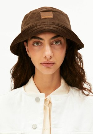 Frau mit langen dunklen Haaren, die einen braunen Bucket Hat mit der Aufschrift „ARMEDANGELS“ trägt, und eine weiße, geknöpfte Jacke über einem cremefarbenen Shirt.
