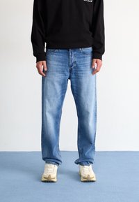 Inimesel on seljas helesinised sirged teksad, valged spordijalatsid ja must pika varrukaga särk, seistes sinisel põrandal.