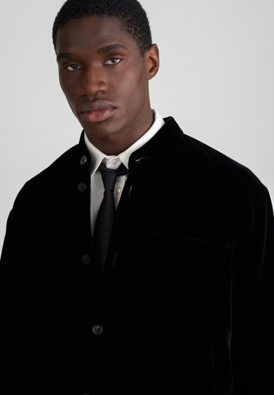 Black velvet jacket with a collar, button closure, and a chest pocket, worn over a white shirt and black tie. Smooth texture.