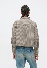 Osoba z warkoczami, ubrana w brązowo-białą pasiastą koszulę z długim rękawem i wyblakłe niebieskie dżinsy, zwrócona tyłem na jednolitym tle.