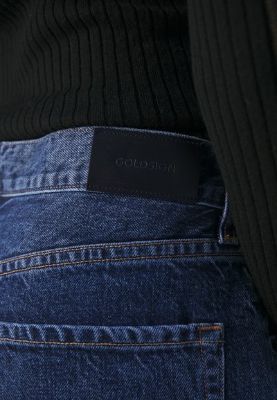 Jean en denim bleu avec une �étiquette en cuir noir cousue sur la taille portant l'inscription "GOLDSIGN". Le tissu a une texture classique avec un léger éclat.