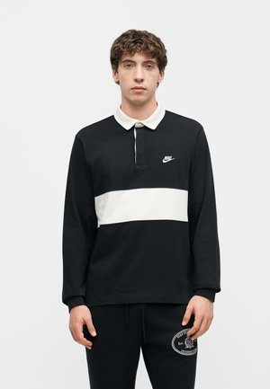 Mladý muž oblečený v čierno-bielej dlhoruká Nike polokošeli a čiernych teplákoch Nike, stojaci pred jednoliatym svetlým pozadím.