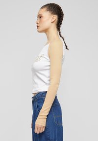 Hvit crop topp med beige lange ermer og et gyllent grafisk design. Kombinert med blå høyhalsa jeans med lomme detaljer.