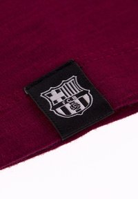 Bordeaux stof met een zwart geweven label met een wit wapenschild ontwerp. Het label toont strepen en initialen, wat de merkidentiteit aangeeft.