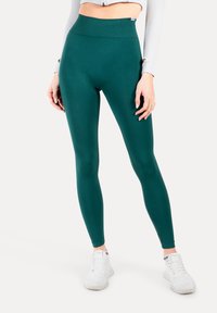 Leggings verdi a vita alta realizzati in materiale elastico. Presentano una texture liscia e un design affusolato, abbinati a scarpe da ginnastica bianche.