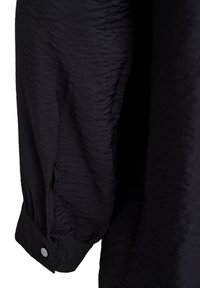 Polsino di una camicia nera a maniche lunghe con un singolo bottone d'argento, tessuto strutturato e leggero dettaglio a piega vicino al polso.