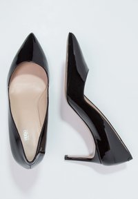 Lesklé černé kožené dámské boty na vysokém podpatku s špičatou špičkou, jedna bota směřující nahoru a druhá ukazující boční profil.