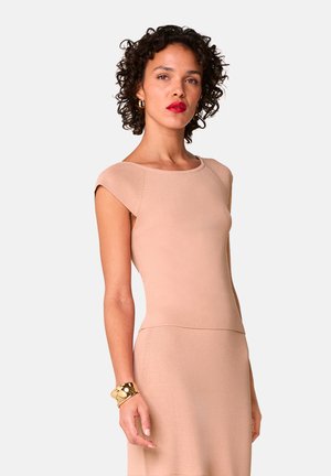 Knielang, getailleerde jurk van zachte beige stof met een ronde halslijn en kapmouwen. Heeft een gladde textuur en strakke lijnen. Gouden bangle om de pols.