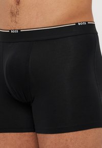 Schwarze Boxershorts aus glattem Stoff mit einem elastischen Bund, der mit Streifen in Kontrastfarbe versehen ist. Das Design ist enganliegend und verfügt über eine konturierte Tasche.