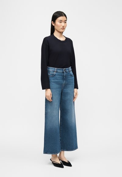 Γυναίκα όρθια που φοράει μαύρο μακρυμάνικο τοπ, μπλε τζιν παντελόνι με φαρδιά μπατζάκια και μαύρα γόνατα με μυτερή μύτη και λουράκι στον αστράγαλο, σε λευκό φόντο.