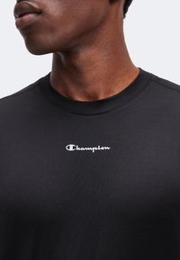 Gros plan d'une personne portant un t-shirt noir Champion avec le logo Champion blanc centré sur la poitrine.