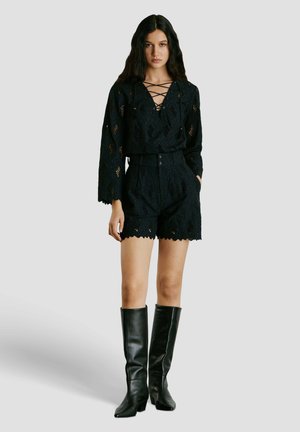 Žena stojaca, oblečená v čiernom krajkovanom top s dlhým rukávom a šnurovacím výstrihom, zladené vysoké šortky a čierne kožené čižmy po kolená.