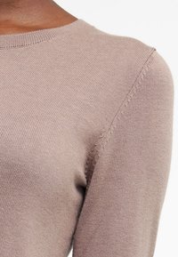 Ljusbrun ribbad stickad tröja med rund hals och synliga sömdetaljer. Slät textur med en figurnära silhuett och långa ärmar.