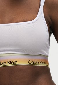 Κοντινό πλάνο σκοτεινόχρωμου ατόμου που φοράει λευκό αθλητικό σουτιέν με λάστιχο Calvin Klein σε χρώματα ουράνιου τόξου.