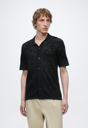 Giovane uomo con capelli mossi che indossa una camicia nera a maniche corte con bottoni e pantaloni beige, in piedi davanti a uno sfondo bianco semplice.
