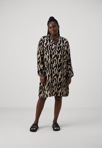Robe à manches courtes, longueur genou en beige avec de grands motifs abstraits noirs ; tissu doux ; détail boutonné au col ; portée avec des sandales noires.