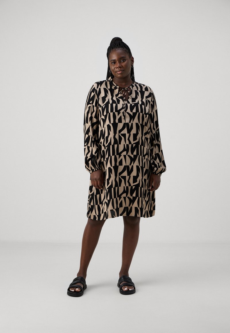 Robe à manches courtes, longueur genou en beige avec de grands motifs abstraits noirs ; tissu doux ; détail boutonné au col ; portée avec des sandales noires.