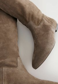 Botas de ante marrón sobre la rodilla con punta afilada, costuras en contraste y un tacón bajo en bloque. Textura suave con un ligero brillo.