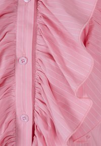 Różowa bluzka z falbankami w białe pionowe paski, z zapięciem na guziki z przodu i teksturowanym materiałem. Miękki, lekki materiał z subtelnymi detalami.