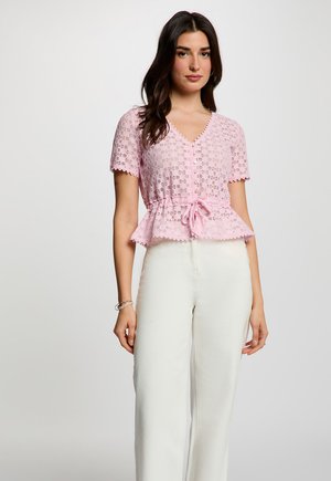 Frau trägt eine rosa Spitzenbluse mit kurzen Ärmeln, Knöpfen und Kordelzug in der Taille, kombiniert mit hoch taillierten weißen Hosen, steht vor einem einfarbigen Hintergrund.