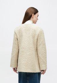 Vrouw draagt een crèmekleurig fleecejack met textuur over een witte coltrui en een blauwe spijkerrok, van achteren gezien tegen een effen achtergrond.