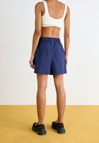 Persona vestida con un top corto blanco, pantalones cortos de cintura elástica azul marino y zapatos deportivos negros, de pie sobre un suelo amarillo contra una pared blanca.