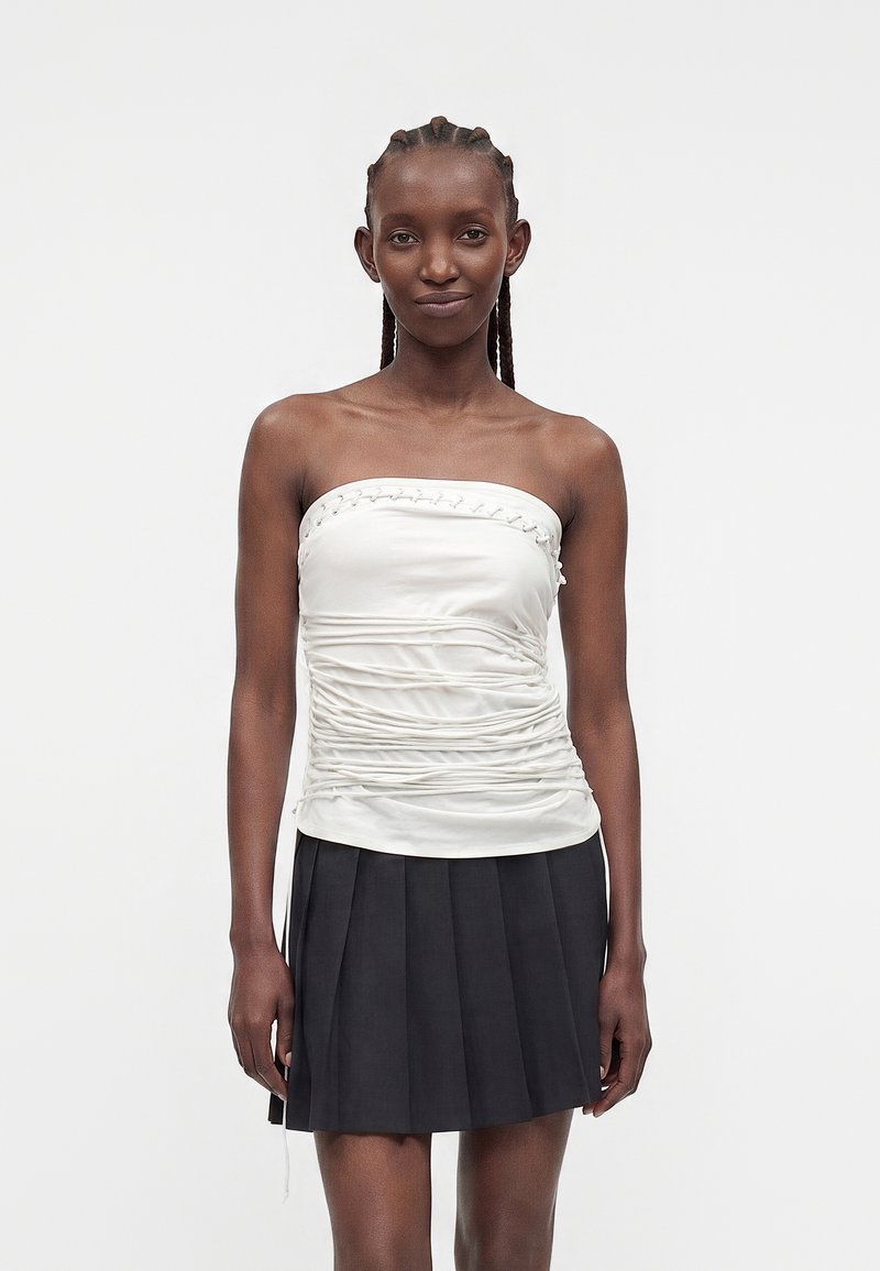 Femme aux cheveux tressés portant un haut bustier blanc froncé et une jupe noire plissée, debout devant un fond clair uni.