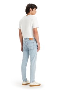 Light blue denim jeans with a tapered fit, featuring a brown leather patch at the back, styled with a white short-sleeved shirt.