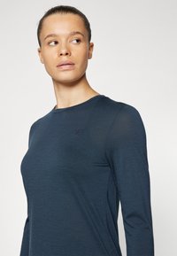 Navy long-sleeve top made from soft, lightweight fabric with a round neckline and subtle logo on the chest. Smooth texture and fitted design.