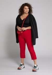 Cardigan noir ouvert, top noir, pantalon rouge cropped et sneakers multicolores. Le modèle tient un petit sac à bandoulière rouge. Cheveux bouclés.